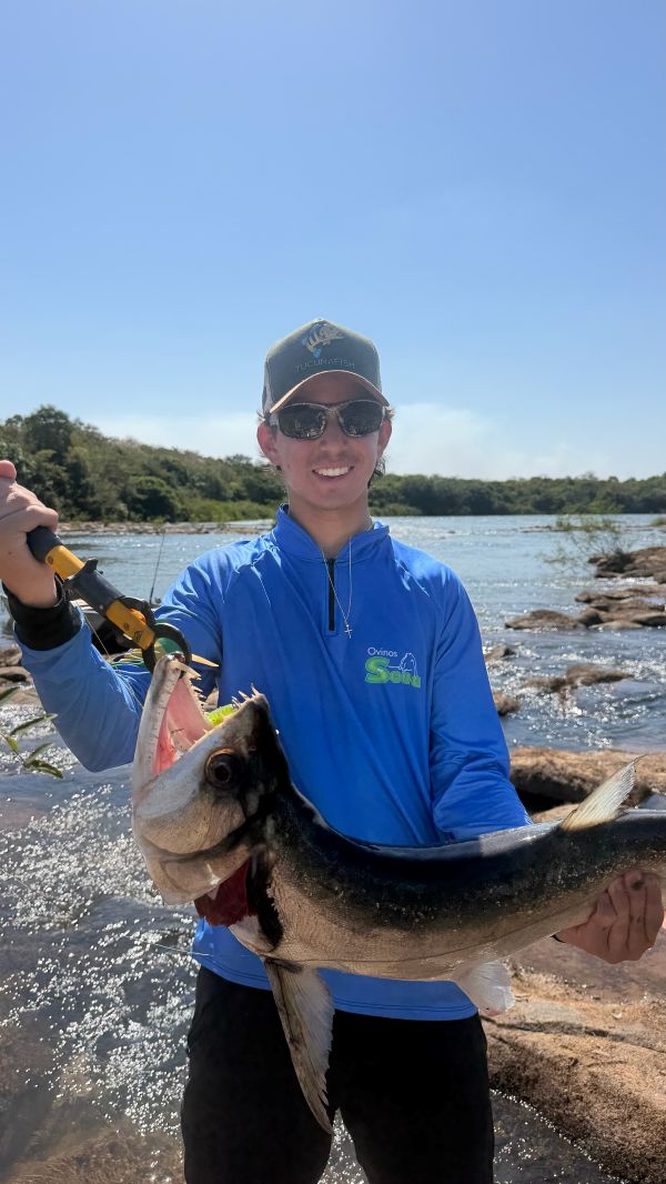 Pescador segurando um peixe no rio, vestindo camisa de pesca TucunaFish azul com zíper, personalizada com logo