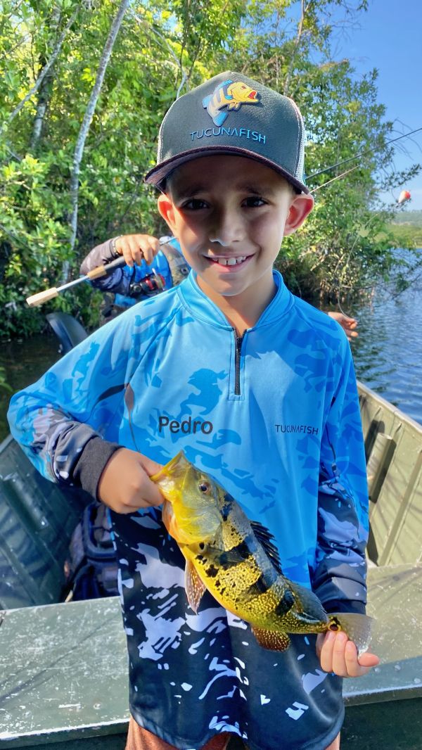 Criança no barco segurando um tucunaré, vestindo camisa de pesca infantil TucunaFish personalizada com nome